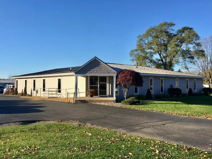 A single-story building with beige siding stands amidst trimmed bushes and trees, under a clear blue sky, on a paved area.